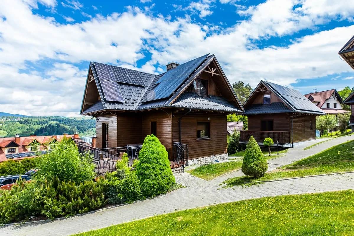 Drewniane domki resortu z ciemnymi dwuspadowymi dachami wśród zieleni, alejka i przystrzyżone krzewy, w tle wzgórza